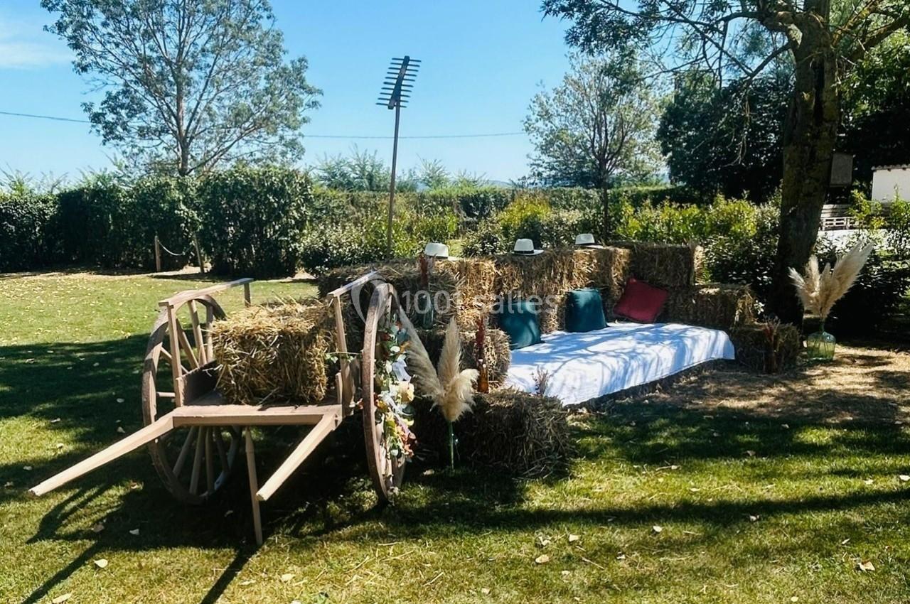 Banc rustique en bottes de paille avec coussins colorés, décor champêtre sous un arbre, près d'une charrette en bois.