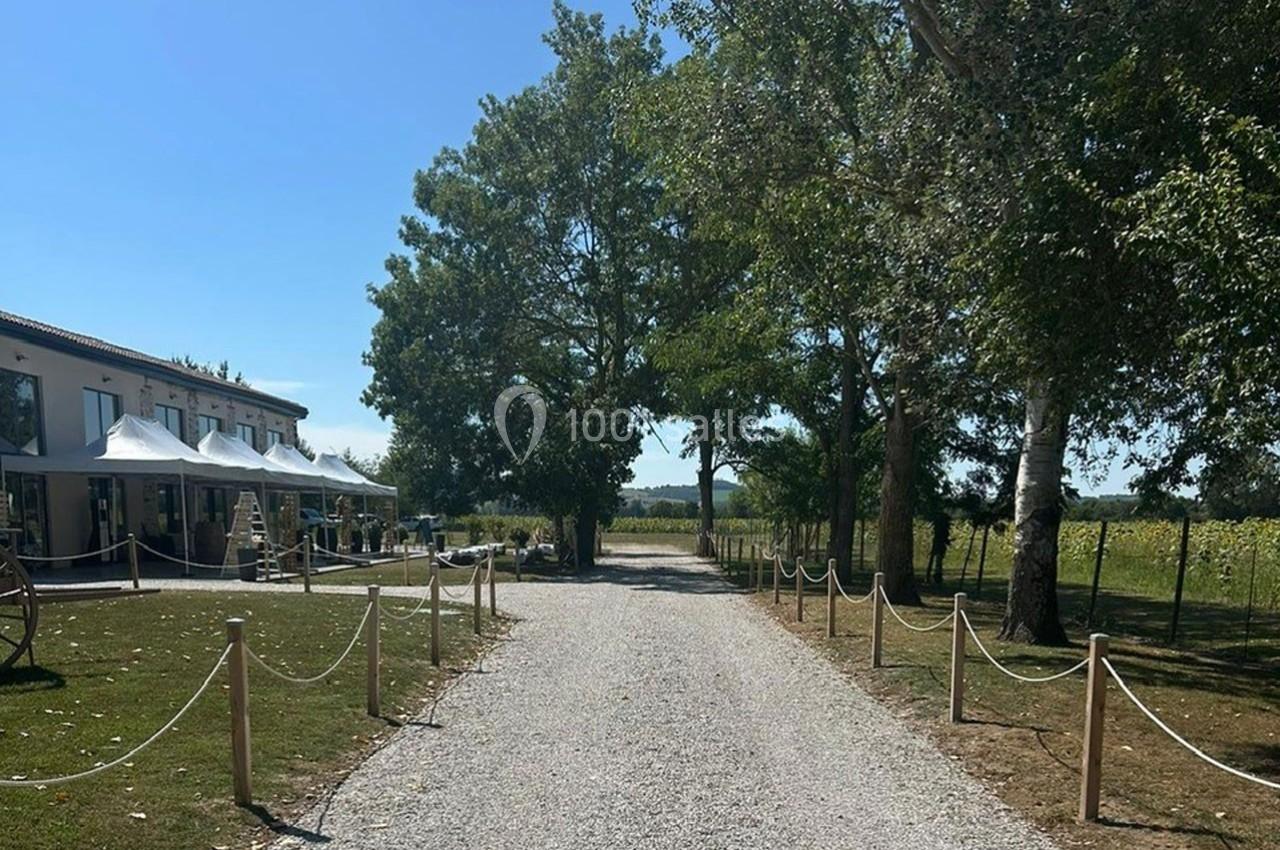 Allée en gravier bordée d'arbres menant à un bâtiment avec des tentes blanches, sous un ciel dégagé.