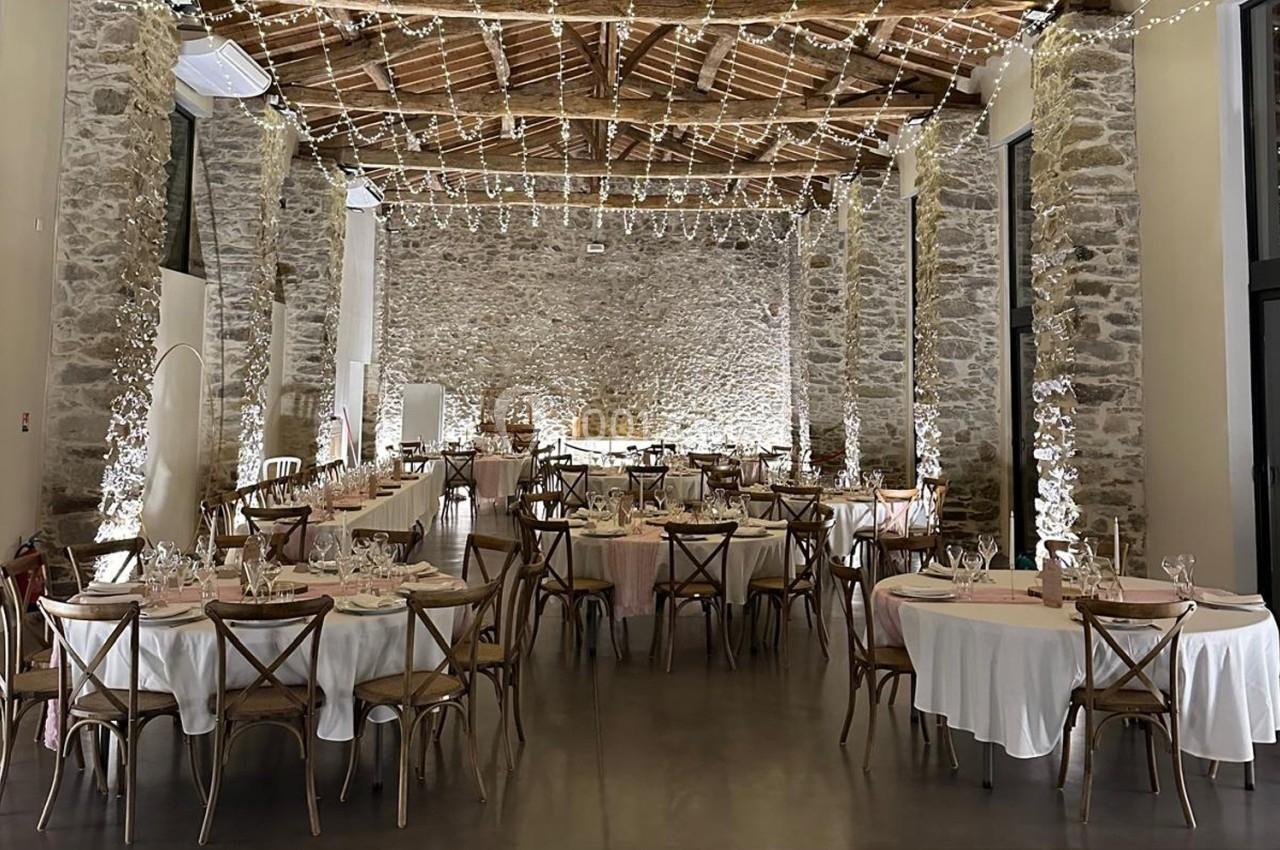 Salle de réception en pierre avec tables rondes dressées, chaises en bois et guirlandes lumineuses suspendues.