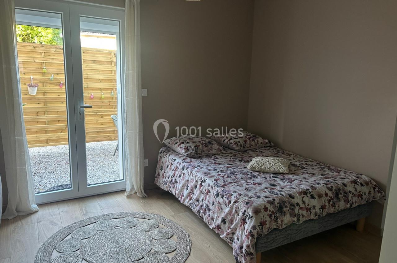 Chambre avec un lit double à motif floral, tapis rond au sol et porte-fenêtre donnant sur une terrasse en bois.