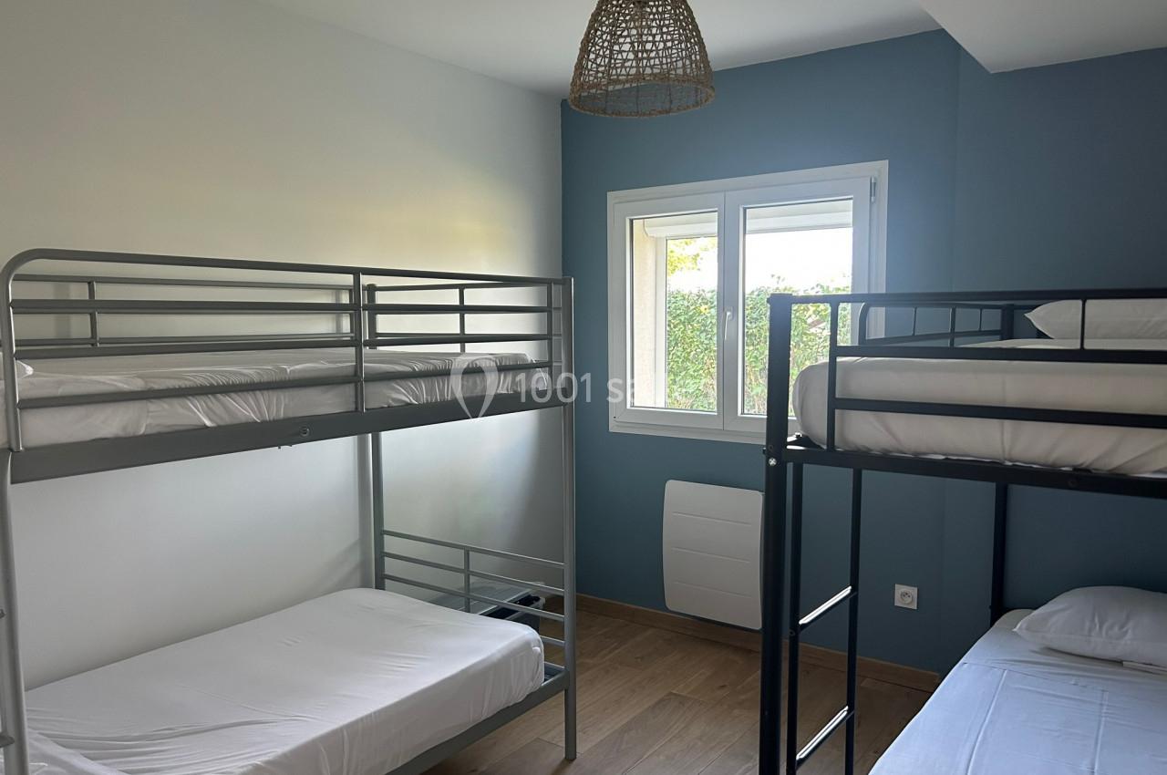 Chambre avec deux lits superposés en métal, murs blancs et bleus, fenêtre donnant sur un jardin, parquet au sol.