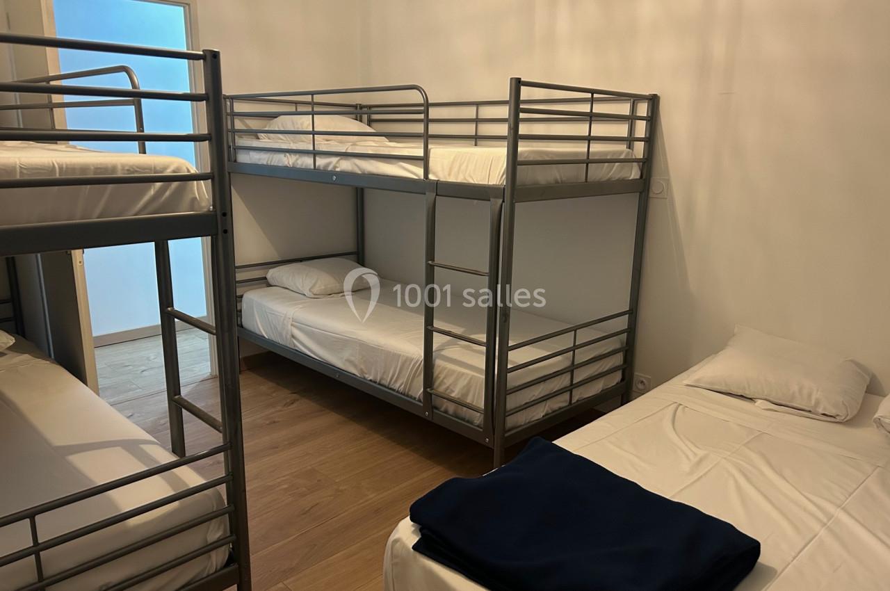 Chambre avec lits superposés en métal, draps blancs et parquet clair, éclairée par une porte vitrée.