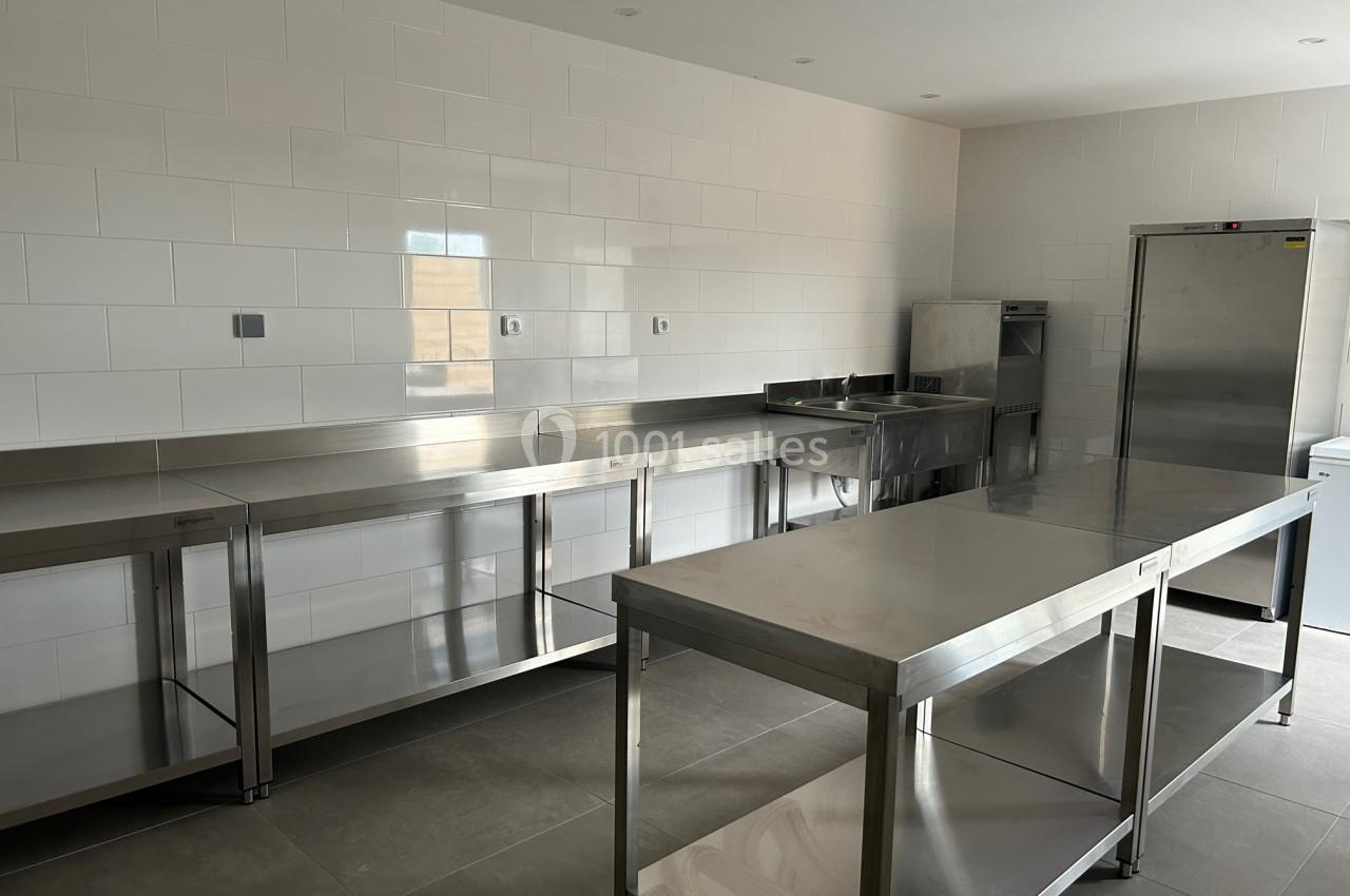 Cuisine professionnelle avec plans de travail en inox, évier, réfrigérateur et murs carrelés blancs.