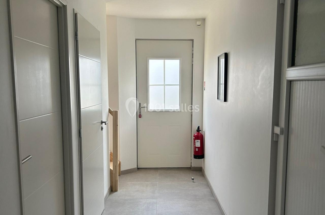 Vue d'un couloir lumineux avec une porte blanche, un extincteur rouge fixé au mur et des portes latérales.