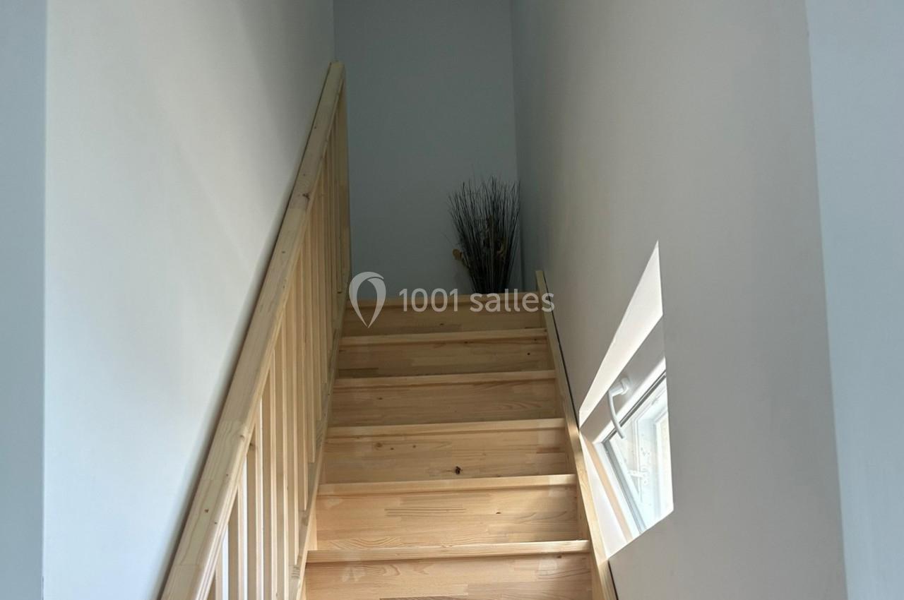Escalier en bois clair avec une rampe en bois, éclairé par une petite fenêtre sur le mur droit.