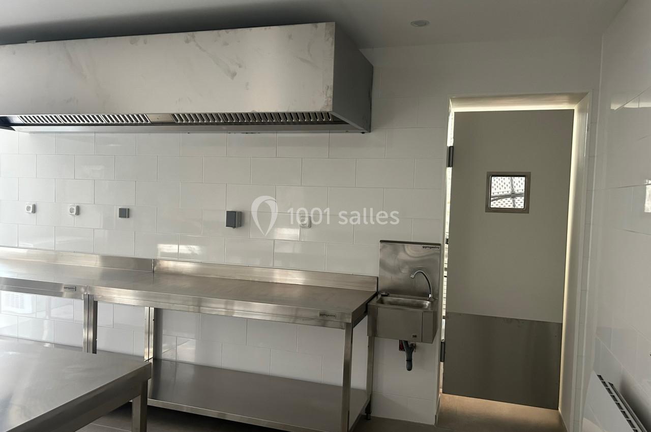 Cuisine professionnelle avec plan de travail en inox, hotte aspirante, évier mural et murs carrelés blancs.