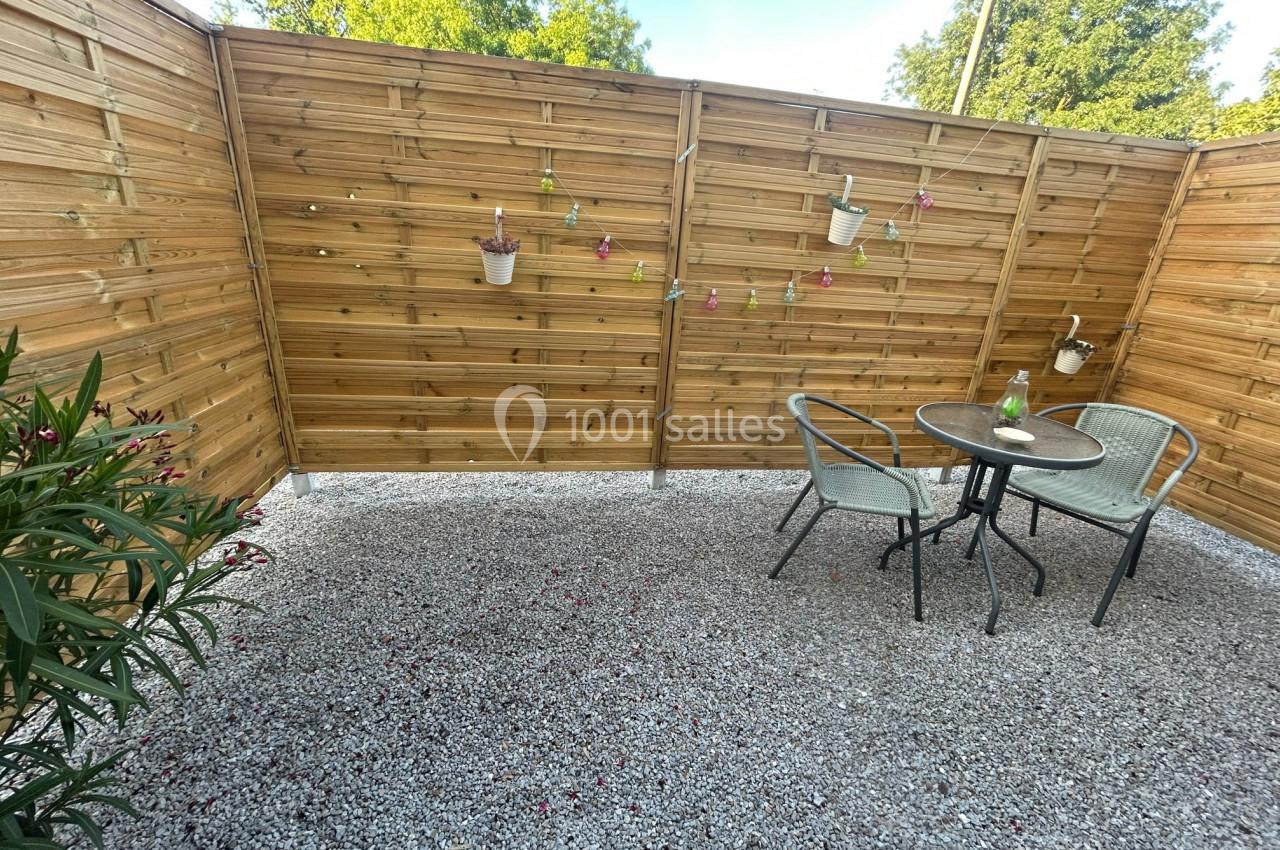 Terrasse en gravier avec table, chaises en métal, clôture en bois décorée de pots suspendus et plantes environnantes.