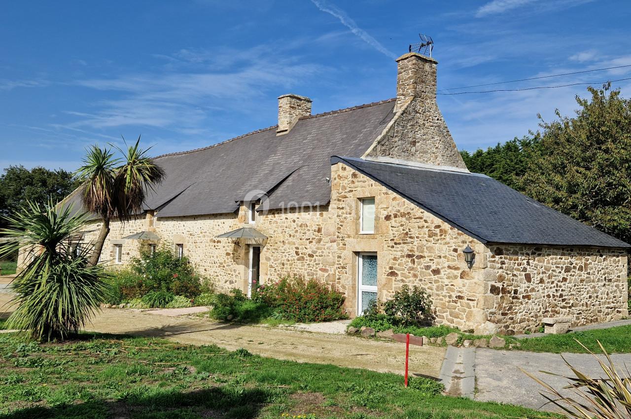 Maison en pierre traditionnelle avec toit en ardoise, entourée de végétation et d'un ciel dégagé.