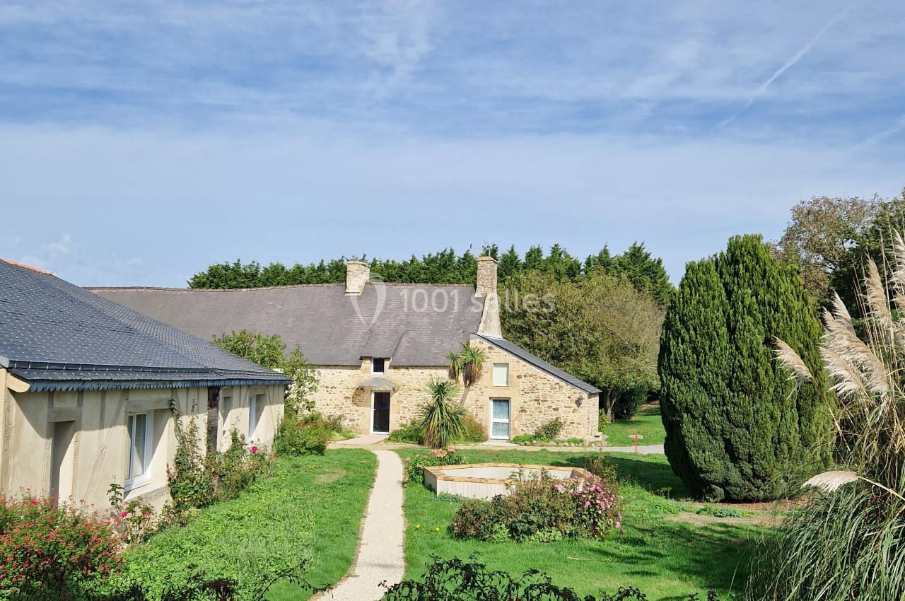 Bâtisse en pierre avec toit en ardoise entourée de verdure, jardin et petit bassin sous un ciel dégagé.