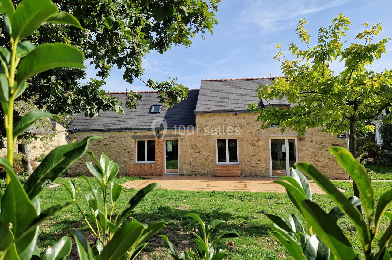 Façade d'une maison en pierre avec toit en ardoise, entourée de verdure et éclairée par un ciel ensoleillé.