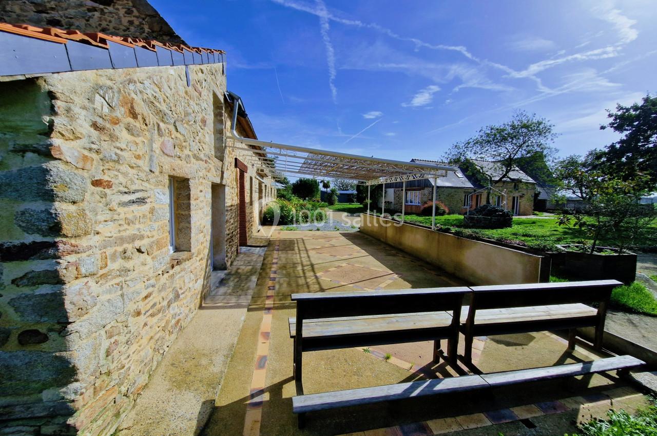 Allée pavée longeant un bâtiment en pierre avec une pergola, bancs en bois et jardin verdoyant sous un ciel bleu.