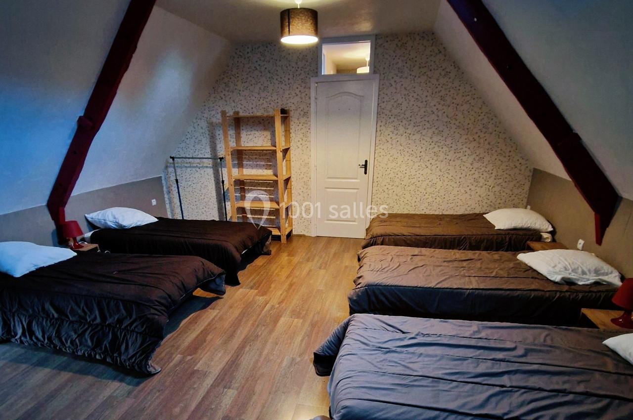 Dortoir mansardé avec six lits simples alignés, parquet au sol, étagère en bois et lumière naturelle d'une fenêtre de toit.