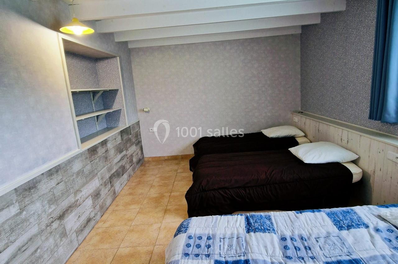 Chambre avec trois lits simples, sol en carrelage, murs clairs, étagères intégrées et plafond avec poutres apparentes.