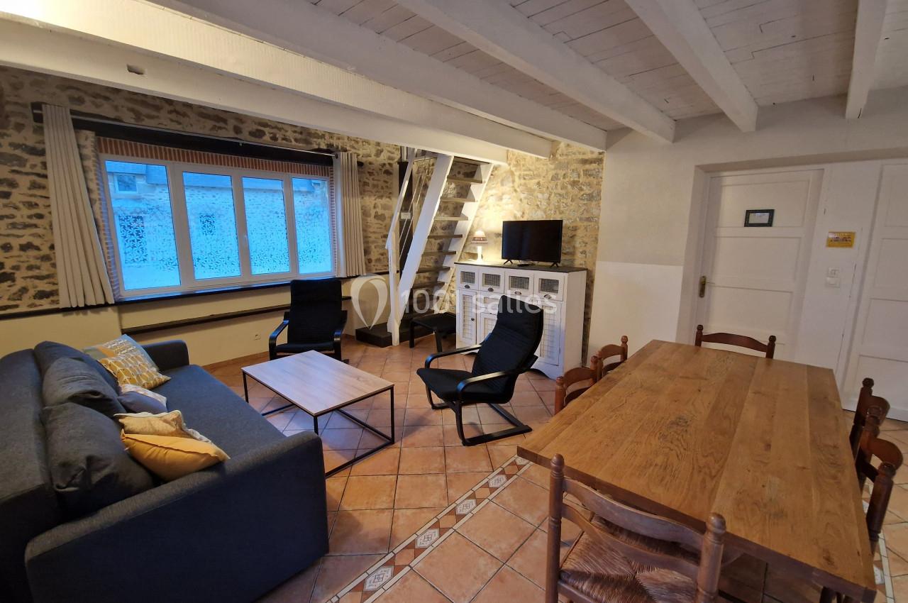 Salon avec murs en pierre, canapé, table en bois, chaises, téléviseur, escalier et grande fenêtre.