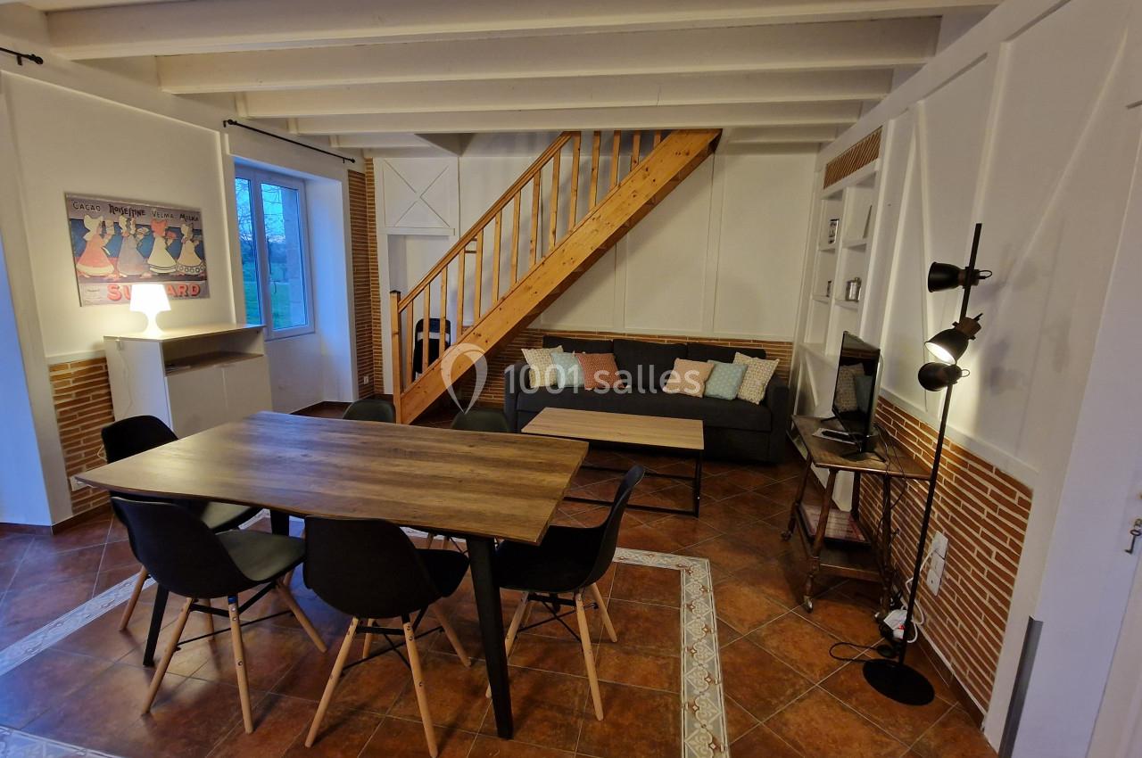 Salle à manger avec table en bois, chaises noires, canapé, escalier en bois et éclairage chaleureux.