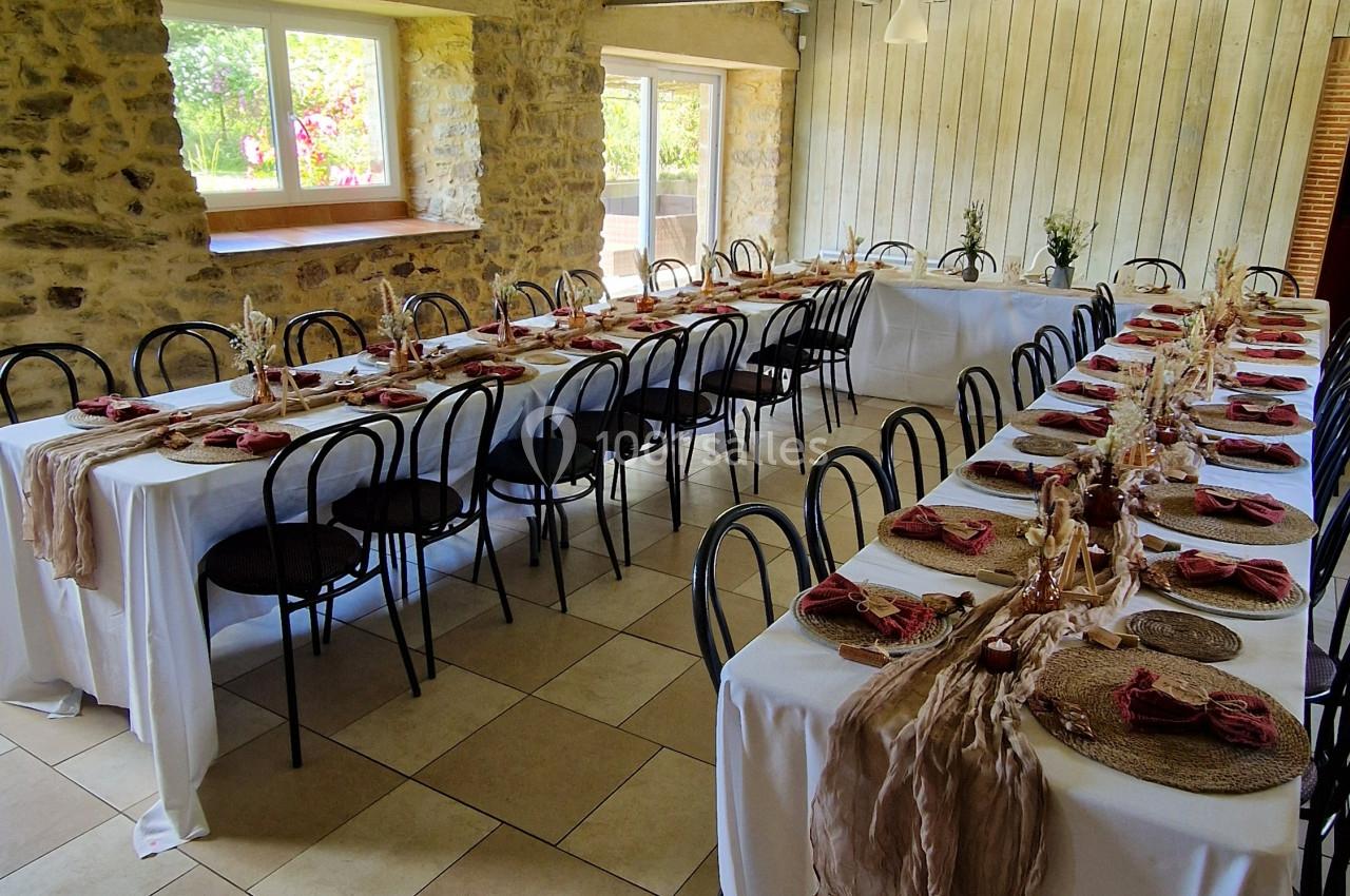 Salle de réception avec tables alignées, nappes blanches, décorations rustiques et lumière naturelle provenant des fenêtres.