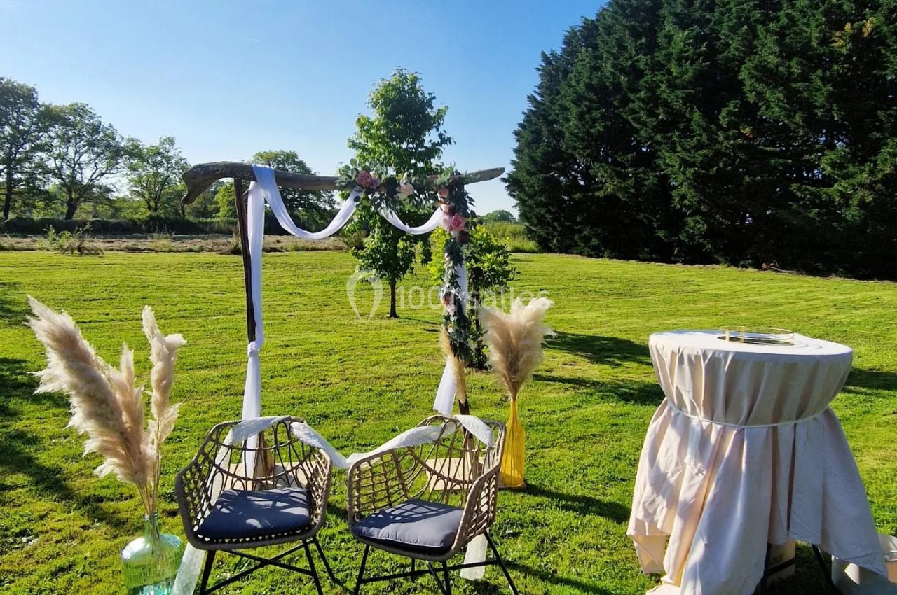 Arche décorée avec des voiles et des fleurs, deux chaises en osier et une table drapée sur une pelouse ensoleillée.