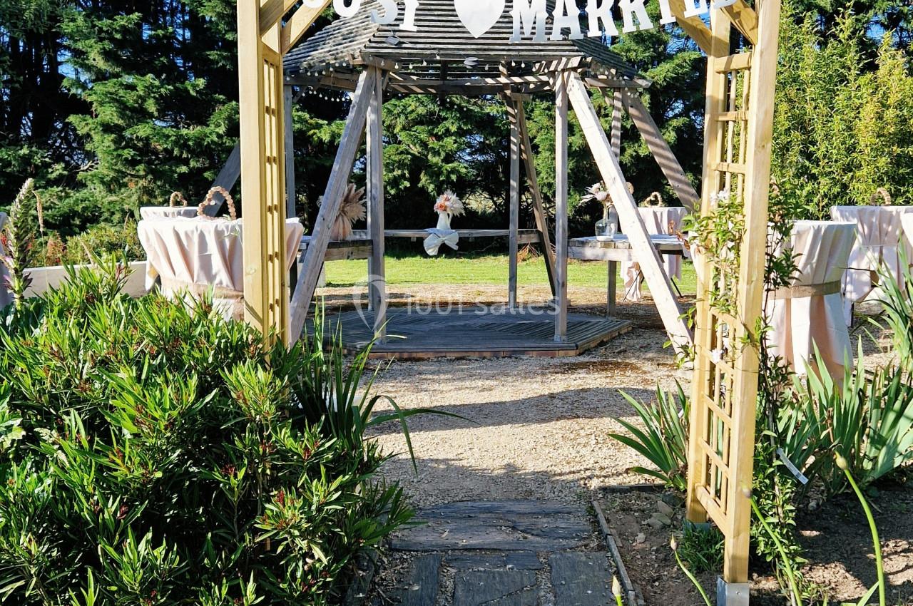 Entrée d'une pergola en bois décorée d'une bannière ’Just Married’, entourée de verdure et de tables dressées.