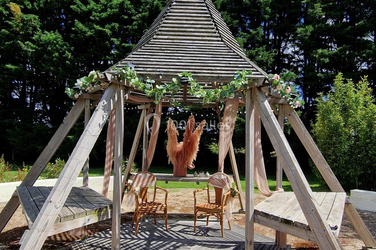 Structure en bois avec toit en pointe, décorée de guirlandes, abritant des chaises sur une terrasse ensoleillée.