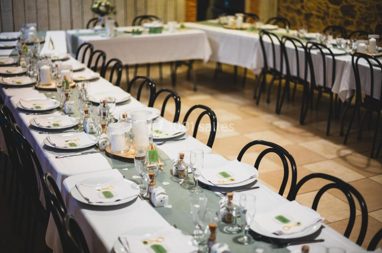 Tables dressées avec nappes blanches, vaisselle et décorations, disposées dans une salle de réception lumineuse.