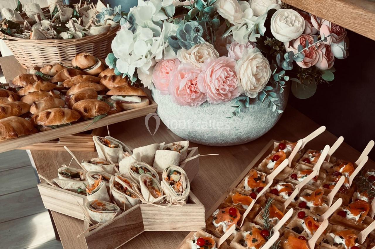 Plateau de bouchées apéritives variées avec wraps, feuilletés et canapés, décoré de fleurs en arrière-plan.