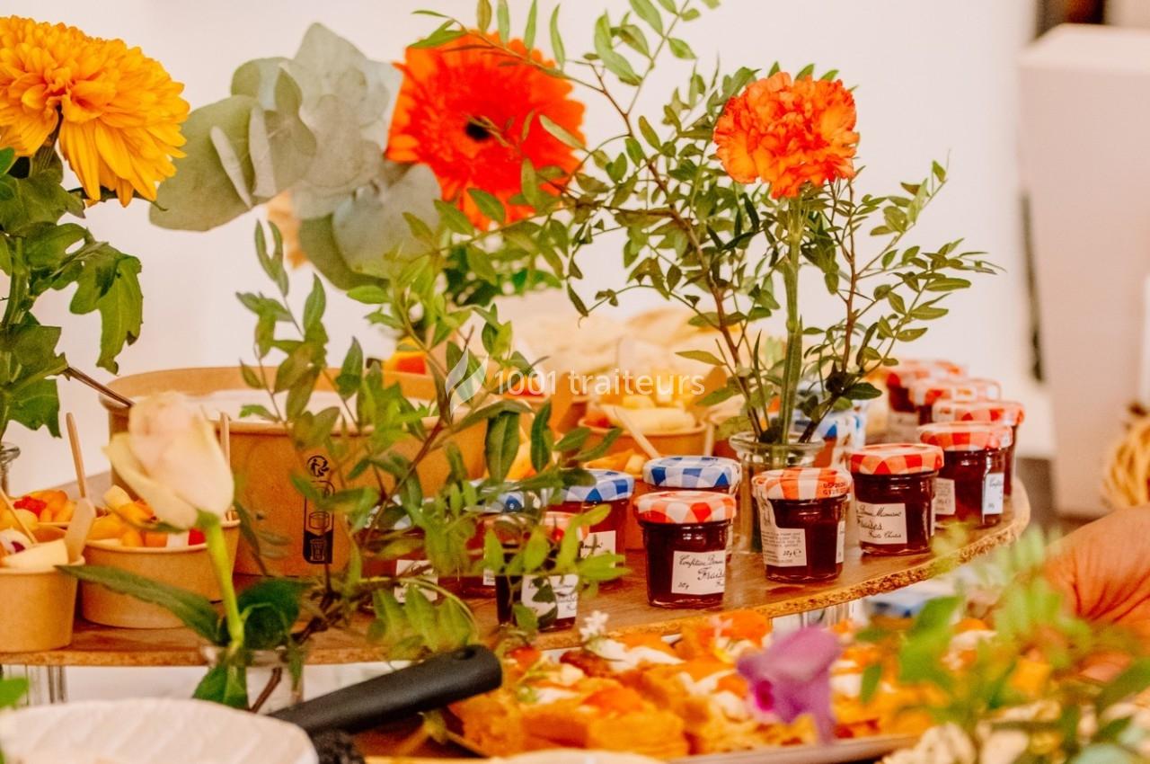 Table garnie de pots de confiture, fleurs colorées et assortiments alimentaires dans un cadre lumineux.