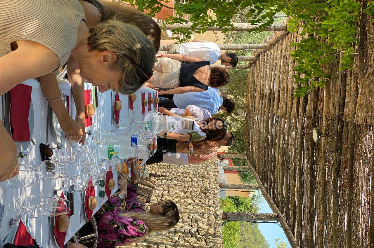 Des convives assis et debout partagent un repas en extérieur sous une pergola ombragée, entourés de verdure.