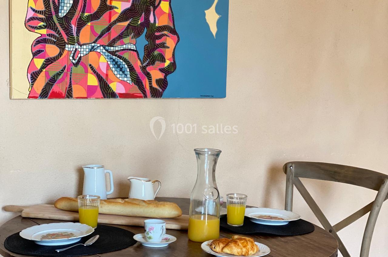 Table en bois avec petit-déjeuner composé de croissants, jus d'orange et café, devant un tableau coloré sur un mur beige.