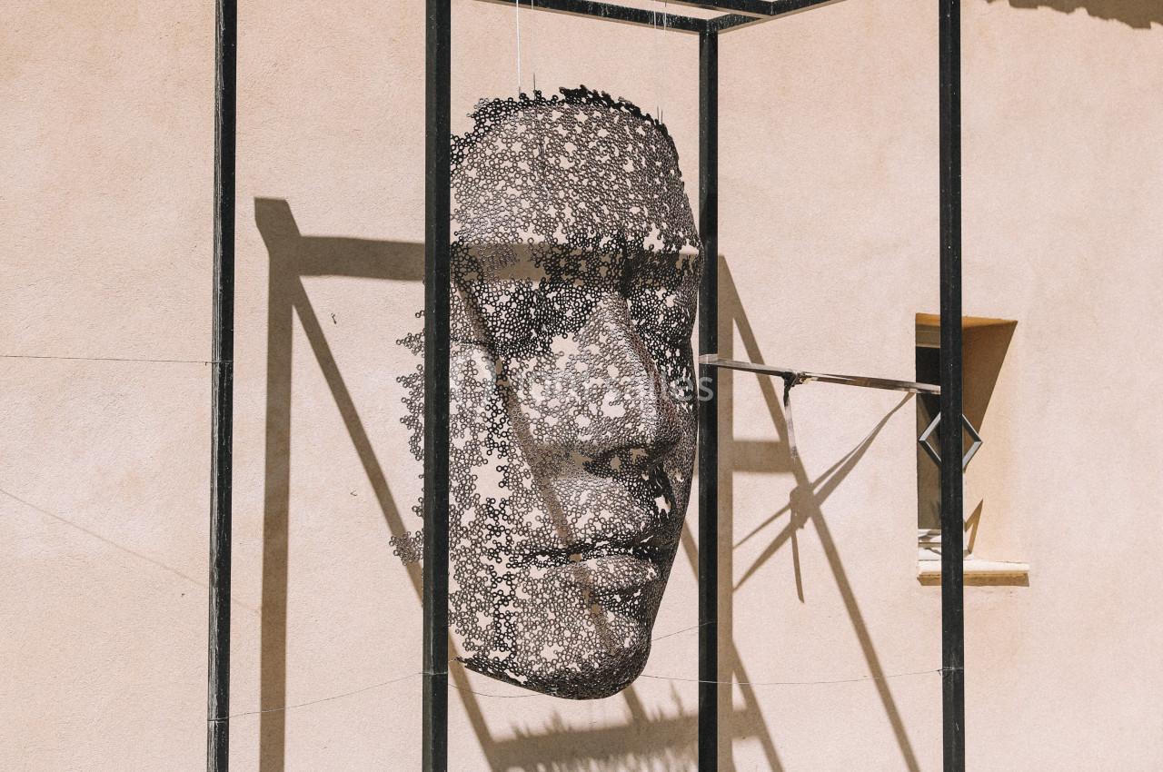 Sculpture suspendue représentant un visage humain, encadrée par une structure métallique, sur une place pavée.