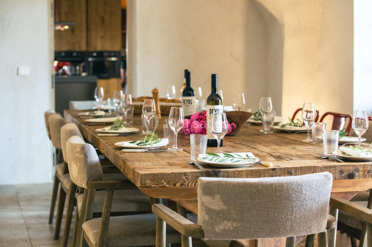 Grande table en bois rustique dressée avec des assiettes, verres et couverts, entourée de chaises rembourrées.