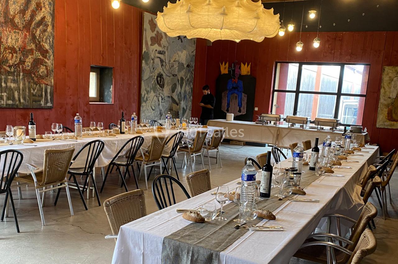 Salle de réception avec tables dressées, nappes blanches, chaises en osier, œuvres d'art et éclairage suspendu.
