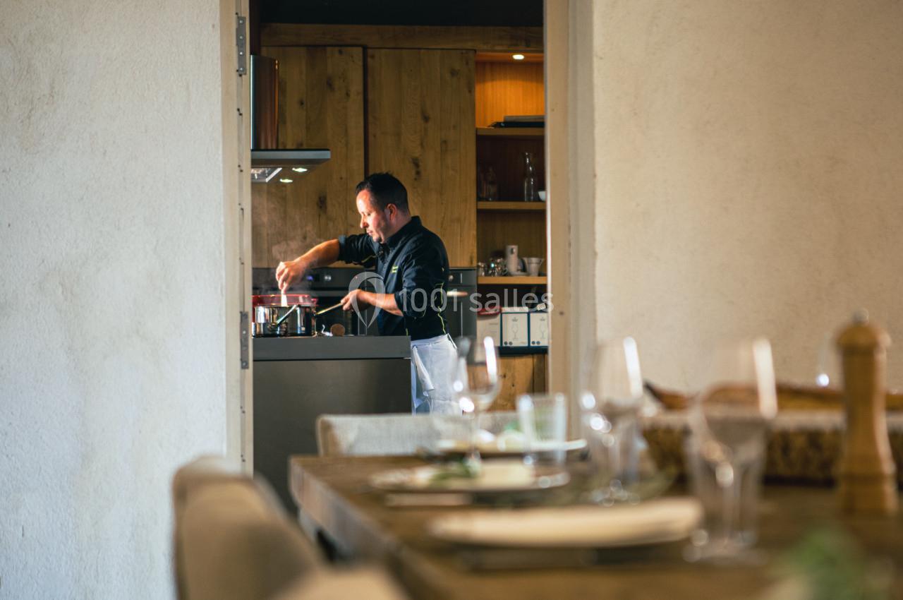 Un chef cuisine dans une cuisine moderne vue depuis une salle à manger avec une table dressée au premier plan.
