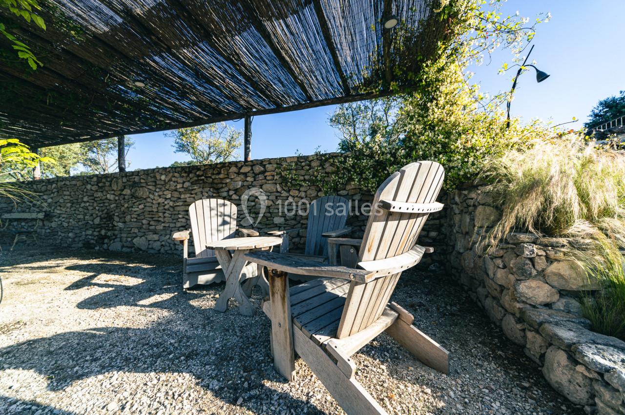 Chaises en bois disposées autour d'une table basse sous une pergola ombragée, avec un mur en pierre en arrière-plan.