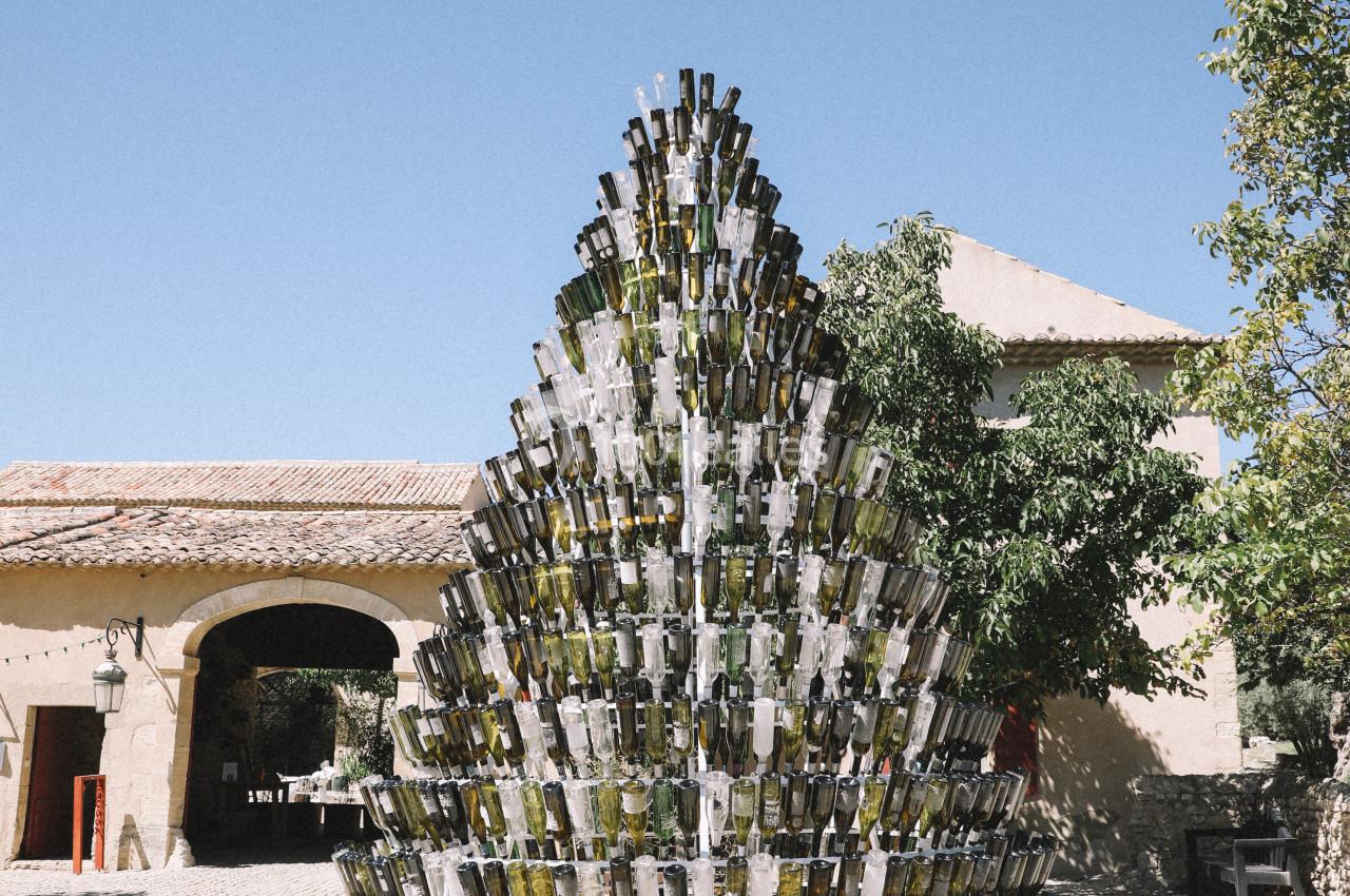 Structure en forme de sapin composée de bouteilles en verre, installée sur une place pavée sous un ciel bleu.