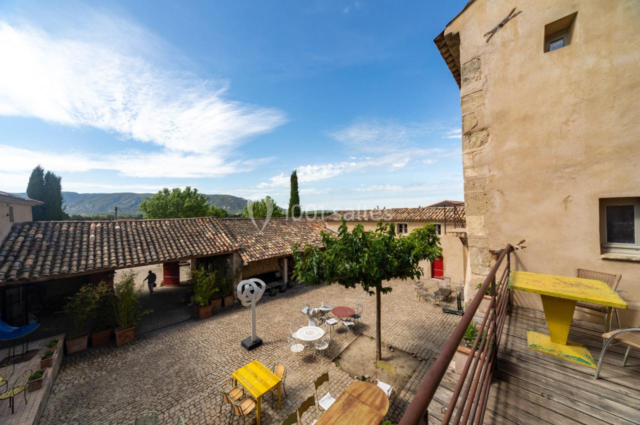 Cour pavée avec des tables extérieures, un arbre central et des bâtiments en pierre sous un ciel dégagé.