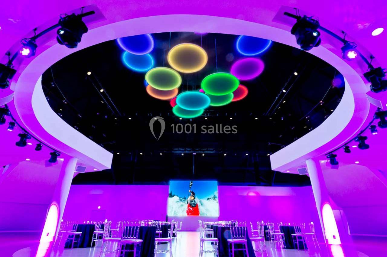 Salle moderne éclairée par des lumières colorées au plafond, avec des tables et des chaises disposées en cercle.