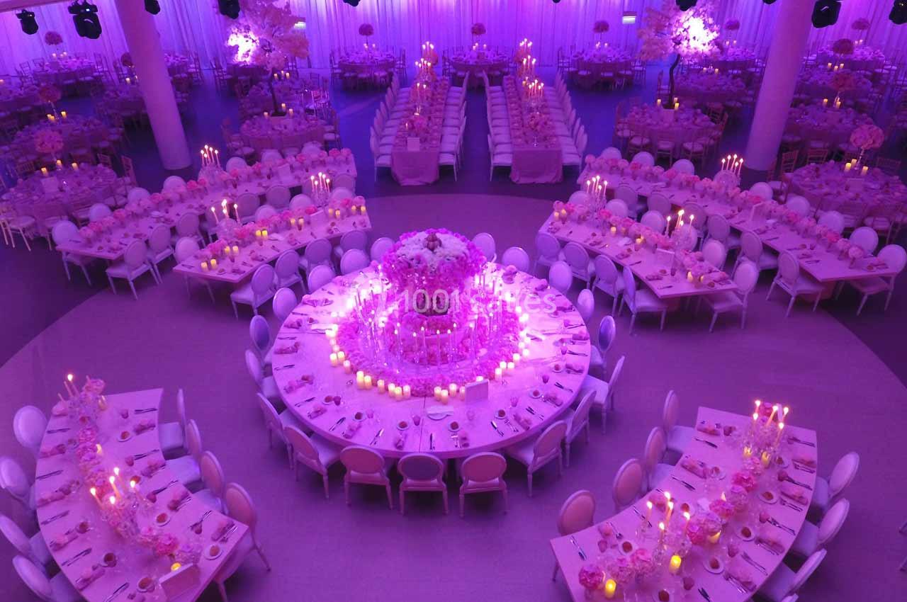 Salle de réception décorée en rose et violet, avec tables rondes et rectangulaires ornées de chandeliers et fleurs.