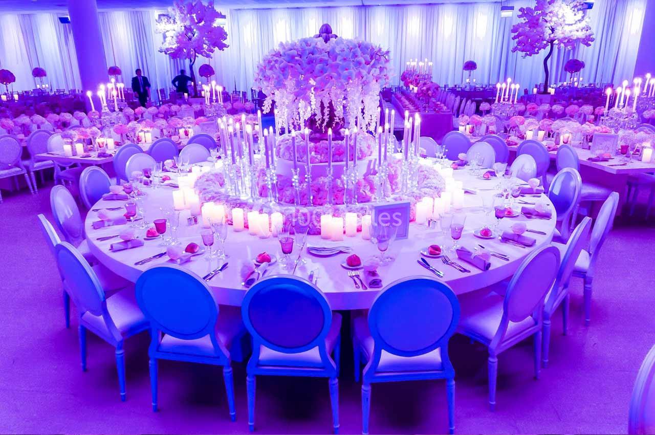 Grande table ronde décorée de fleurs blanches et roses, entourée de chaises bleues, dans une salle éclairée en violet.