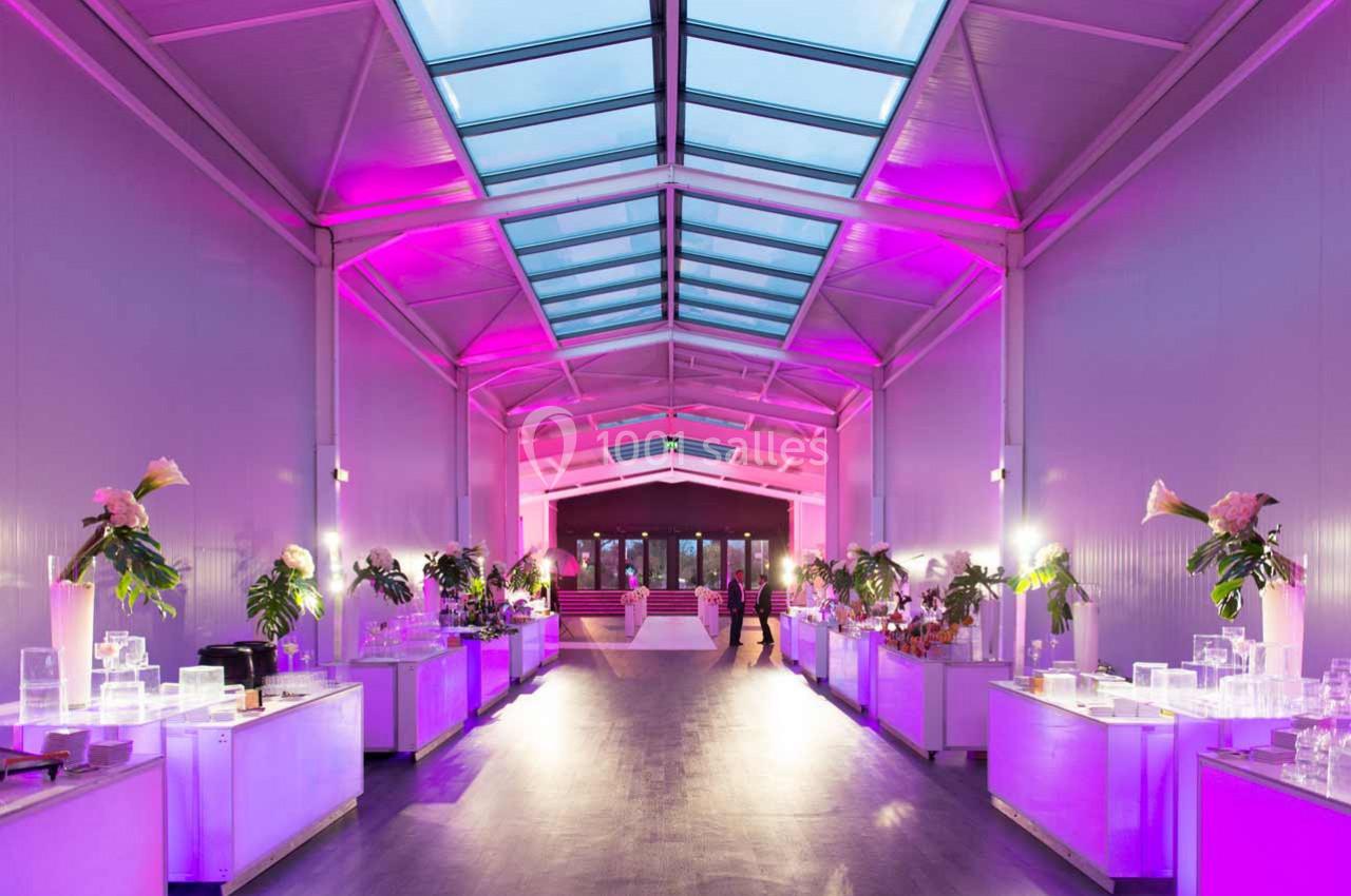 Salle lumineuse avec toit vitré, éclairée par des lumières roses, tables décorées de fleurs et objets transparents.