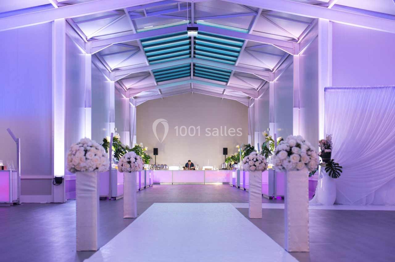 Salle de réception lumineuse avec allée centrale blanche, colonnes fleuries et éclairage violet.