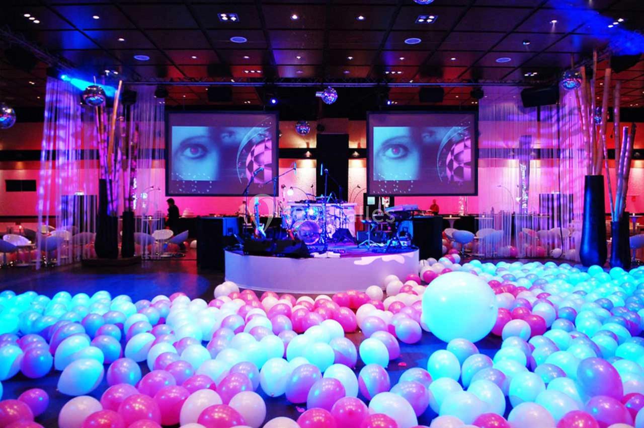 Salle de réception décorée avec des ballons roses et blancs, éclairage coloré et scène équipée pour un événement.