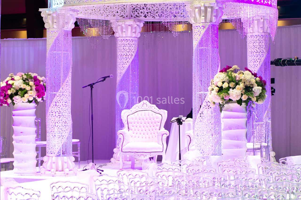 Décor de mariage avec arche ornée de motifs blancs, fauteuil central et arrangements floraux colorés.