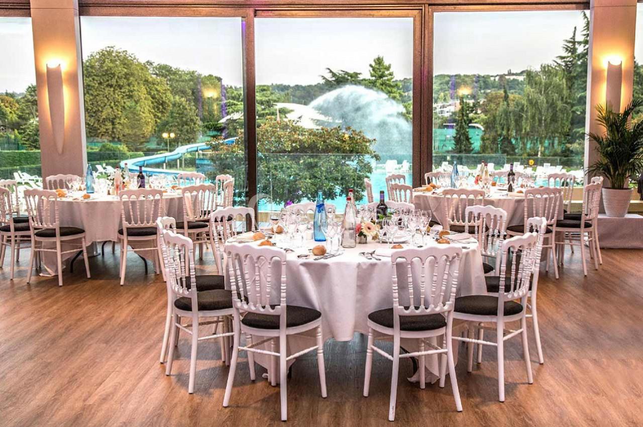 Salle de réception lumineuse avec tables rondes dressées, grandes baies vitrées donnant sur un jardin avec fontaine.