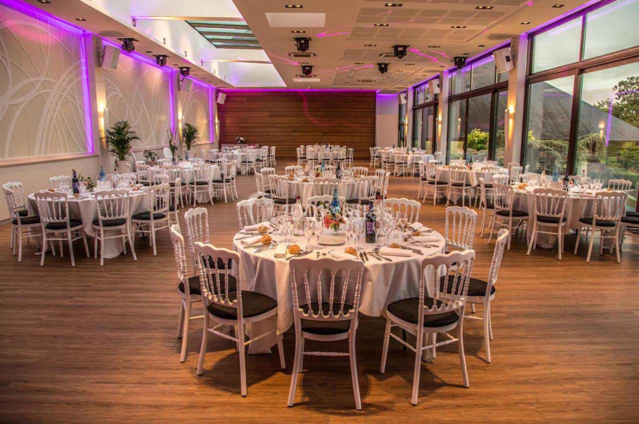 Salle de réception élégante avec tables rondes dressées, chaises blanches et éclairage tamisé.