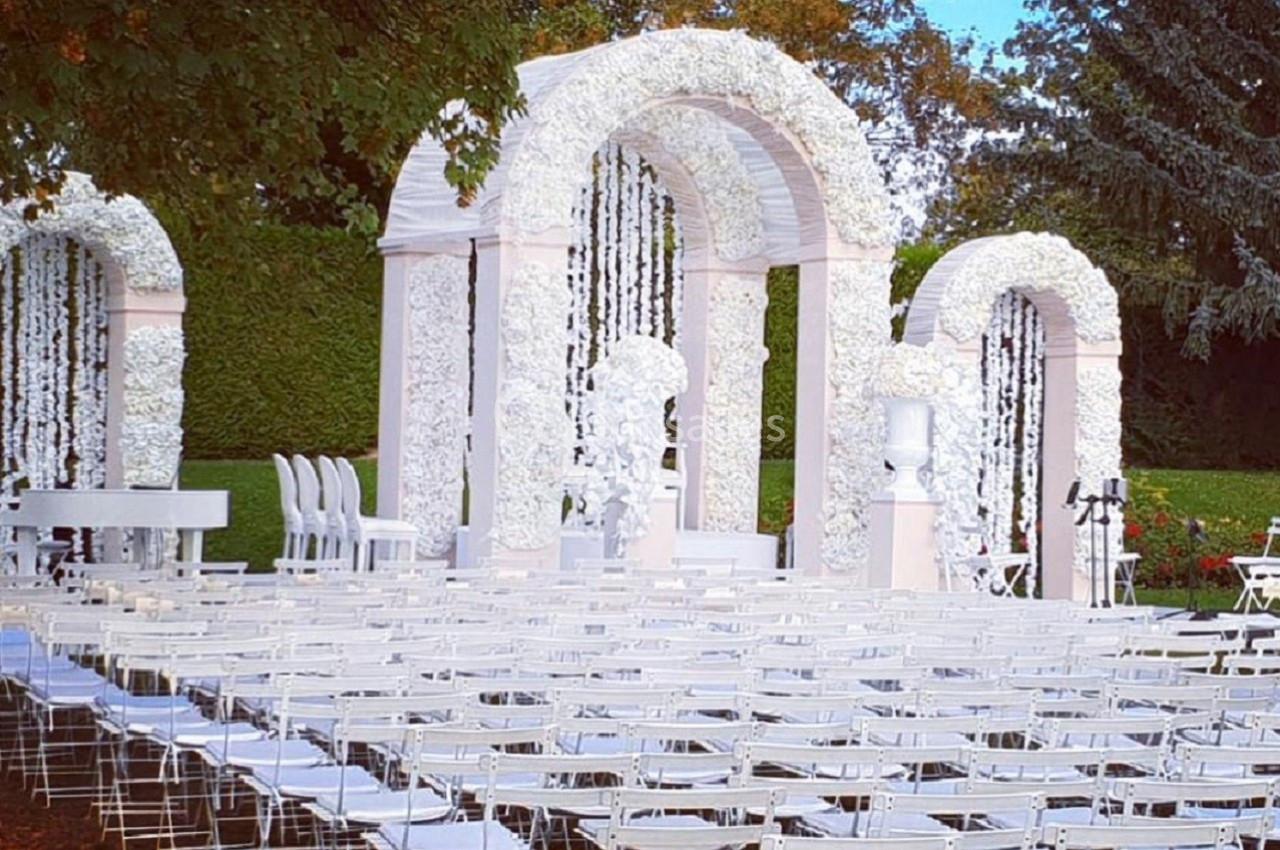 Chaises blanches alignées devant une arche décorée de fleurs blanches dans un jardin pour une cérémonie en plein air.
