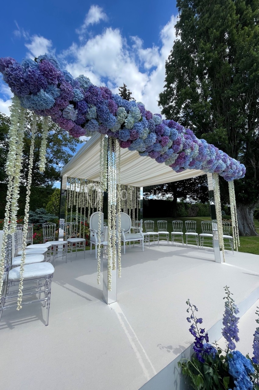 Structure de cérémonie décorée de fleurs violettes et blanches, entourée de chaises, dans un jardin sous un ciel bleu.