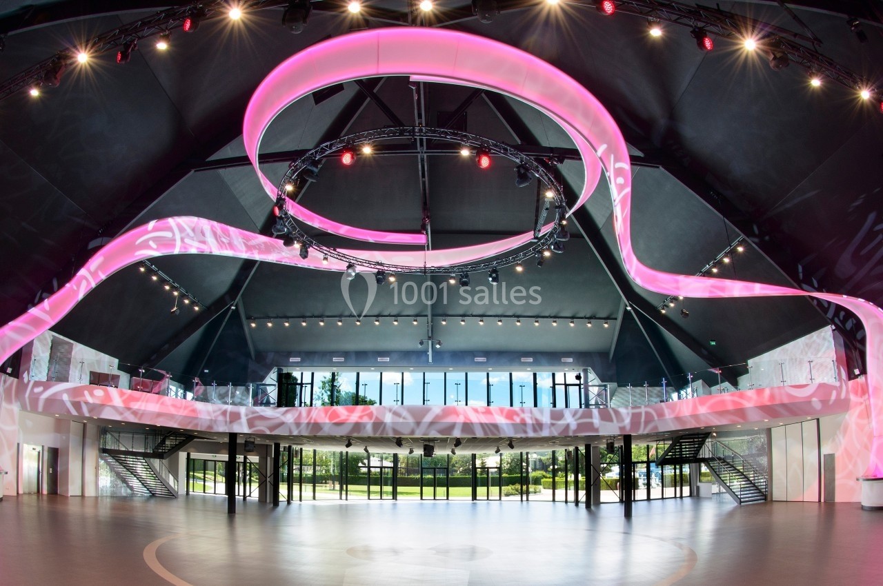 Salle moderne avec un éclairage rose en spirale suspendu, grandes baies vitrées et mezzanine avec escaliers latéraux.