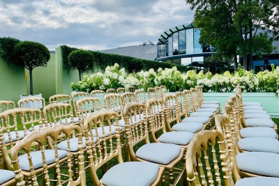 Cérémonie en plein air avec arche florale blanche et rose, chaises transparentes et pelouse verte sous un ciel dégagé.