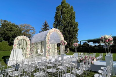 Cérémonie en plein air avec arche florale blanche et rose, chaises transparentes et pelouse verte sous un ciel dégagé.