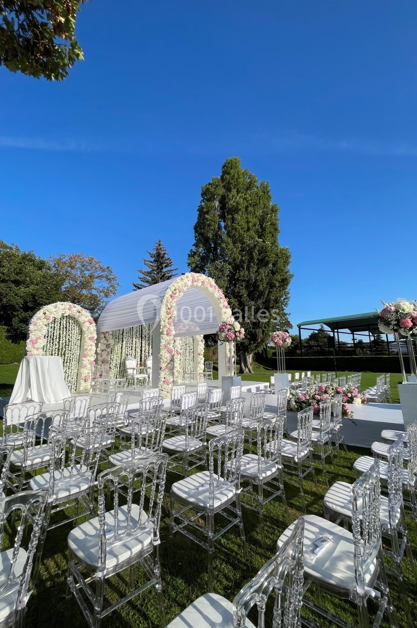 Cérémonie en plein air avec arche florale blanche et rose, chaises transparentes et pelouse verte sous un ciel dégagé.